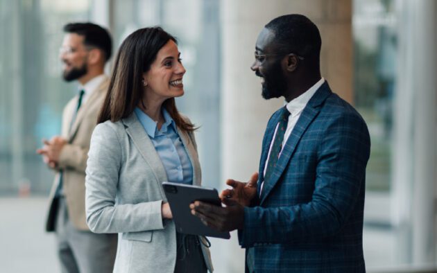 Two business professionals are talking and smiling indoors, with one holding a tablet. Another person stands in the background, looking away.