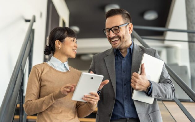 Two people standing on stairs, smiling and talking. One holds a tablet and the other holds a laptop. They appear to be engaged in a business discussion.