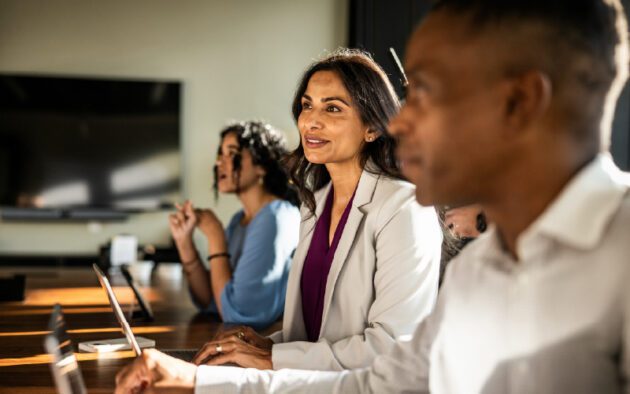 Three people sit at a conference table with laptops, attentively listening during a meeting in a modern office setting.