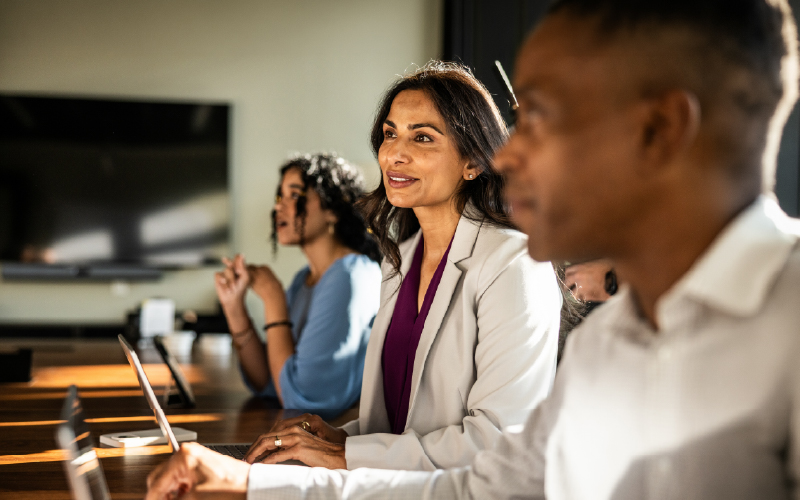 Three people sit at a conference table with laptops, attentively listening during a meeting in a modern office setting.