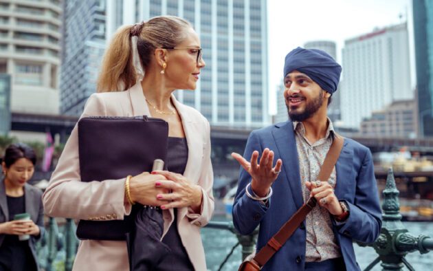 A woman and a man in business attire walk and talk outdoors in an urban setting with tall buildings in the background.