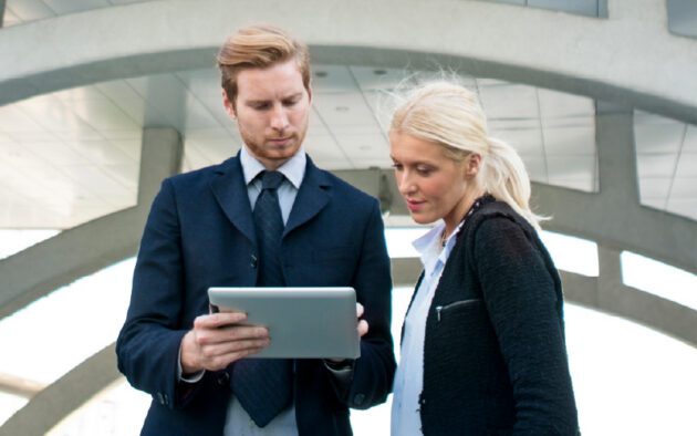 Two business professionals standing outdoors look at a tablet together, with a modern architectural structure in the background.
