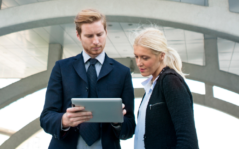 Two business professionals standing outdoors look at a tablet together, with a modern architectural structure in the background.