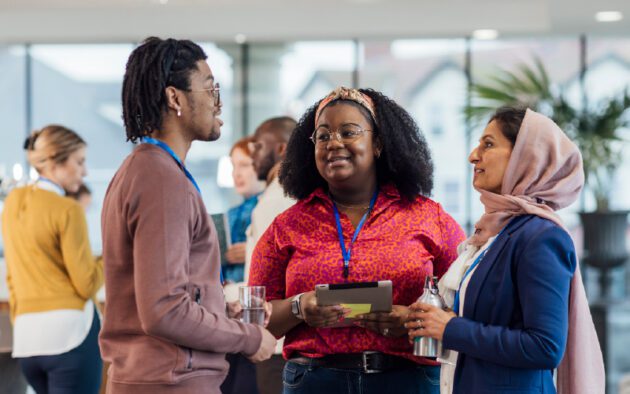 Three people wearing conference badges talk together indoors; two hold drinks and one holds a tablet. Other attendees converse in the background.