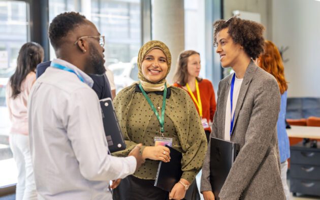 Three people stand together, smiling and talking in a modern office setting, holding laptops. Other professionals mingle in the background.