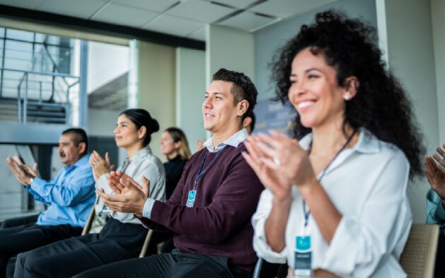 A group of people sit in chairs and clap, appearing attentive and engaged during an indoor event or presentation.