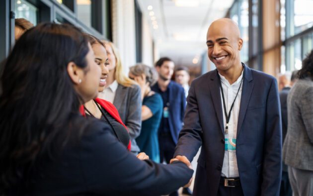 A man in a suit smiles as he shakes hands with a woman at a professional networking event in a brightly lit hallway.