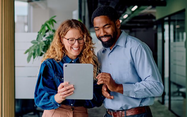 Two people standing indoors look at a tablet together and smile. One holds the tablet while the other points at the screen. Office setting with glass walls and a plant in the background.