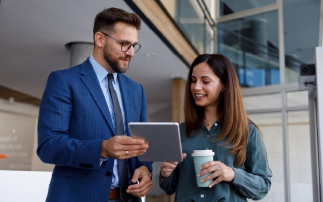 A man in a suit holds a tablet, showing it to a woman holding a coffee cup. They are indoors and appear to be having a discussion.