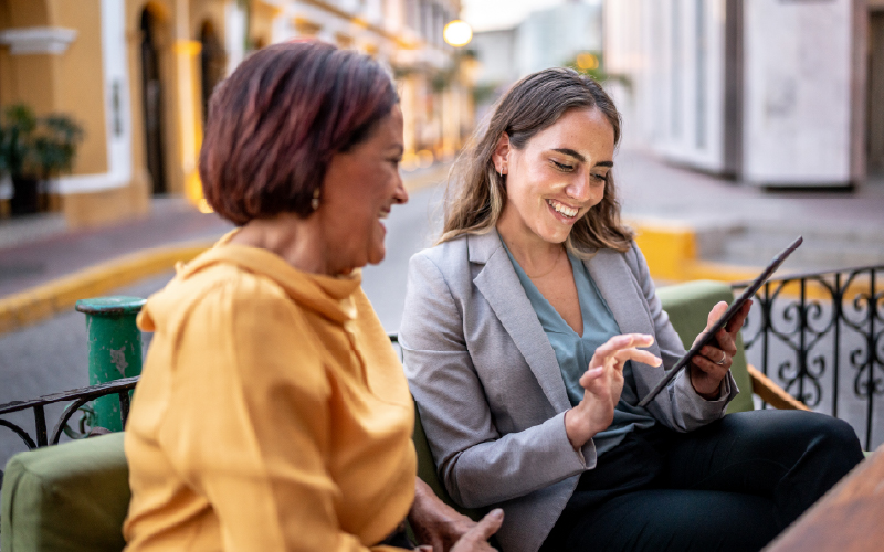 Two women sitting outdoors on a bench, smiling and looking at a tablet together on a city street.