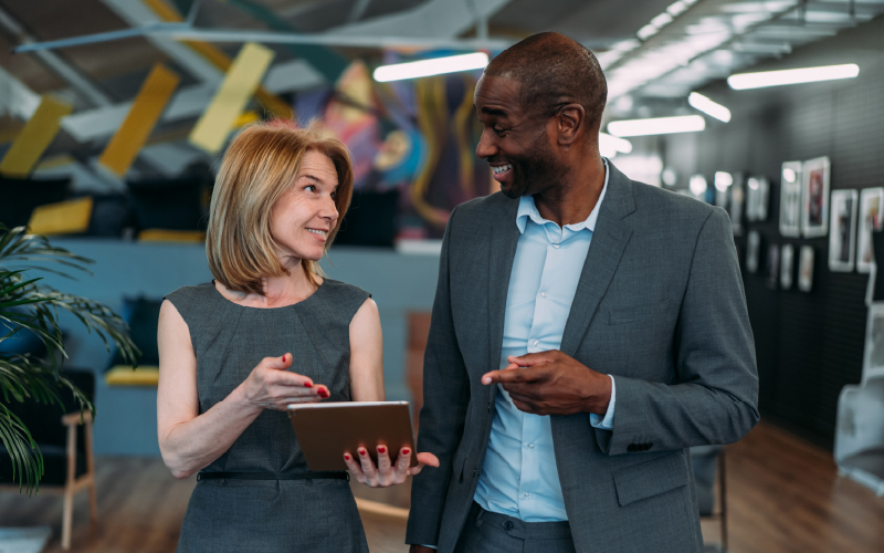 Two business professionals in suits stand indoors, smiling and conversing while one holds a tablet.