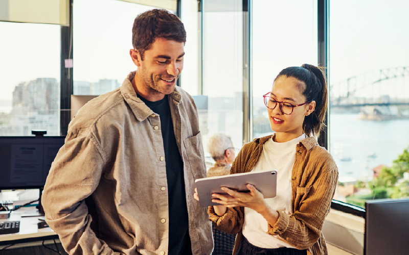 Two people stand in a modern office by a window, looking at a tablet together. A cityscape and bridge are visible outside.