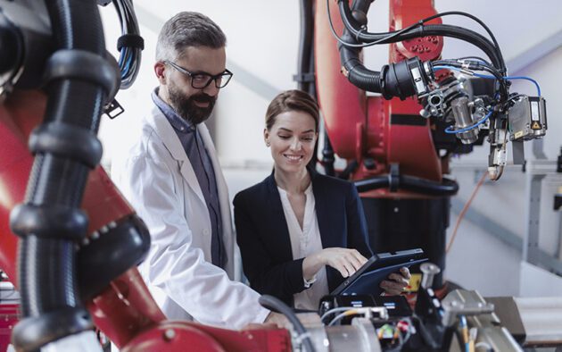 Two professionals, one in a lab coat and one in business attire, examine a tablet and industrial robotic machinery in a modern laboratory or manufacturing facility.