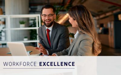 Two professionals in business attire sit at a desk with a laptop, smiling and discussing work. Text overlay reads "Workforce Excellence.