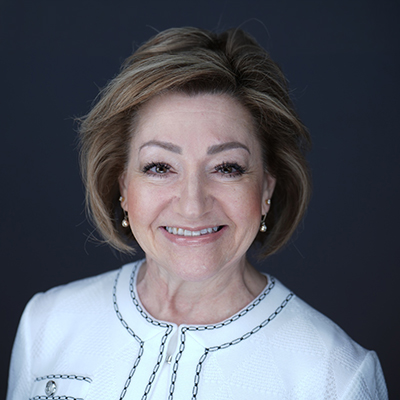 Smiling middle-aged woman with short light brown hair, wearing a white textured jacket with black trim, poses confidently in front of a dark background—an inspiring figure in today’s diverse global workforce.