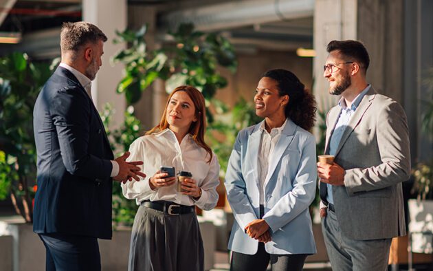 Four professionally dressed people stand and converse in a modern office space with plants and natural light. Two hold coffee cups, one holds a phone.