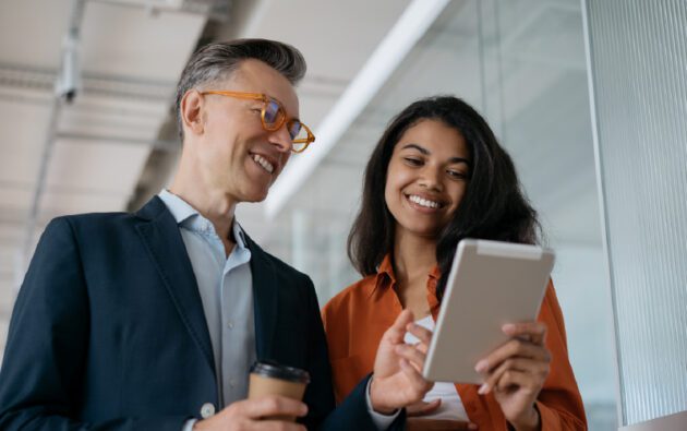 Two people standing indoors, smiling and looking at a tablet. One person is holding a coffee cup. Glass walls and office ceiling are visible in the background.