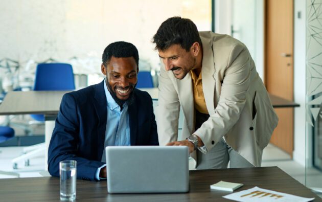 Two men in business attire look at a laptop and smile in a modern office. A glass of water, smartphone, and documents with charts are on the table.