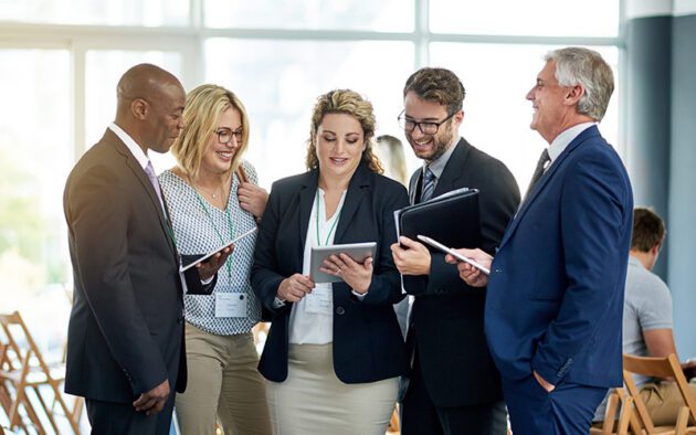 Five business professionals stand in a group, smiling and looking at a tablet and documents during a meeting or conference in a bright room with rows of chairs.