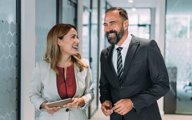Two business professionals, a woman holding a tablet and a man in a suit, are smiling and talking as they walk down an office hallway with glass walls.