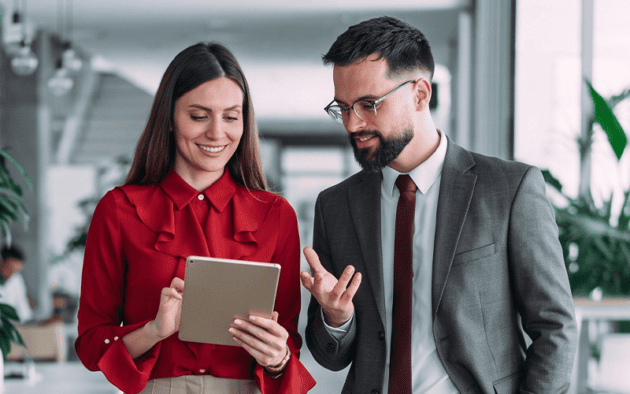 Two professionally dressed people stand indoors, looking at a tablet together and having a discussion about workforce management governance.