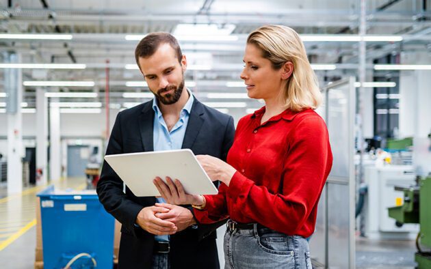 Two people standing in a factory setting, looking at a tablet together. The woman in red is holding the tablet and discussing something with the man in a dark jacket.