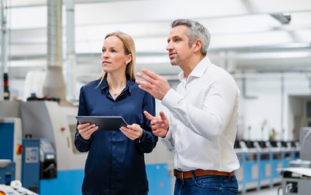 A woman holding a tablet and a man gesturing as they talk in a manufacturing facility.