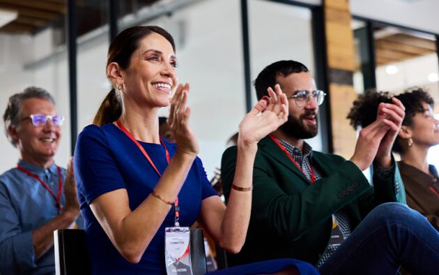 Four people wearing conference badges applaud during a presentation.