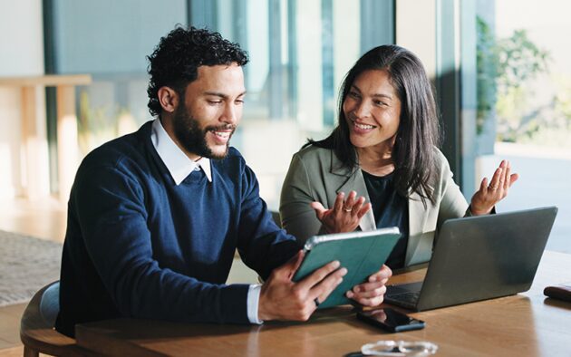 A man and a woman in an office setting look at a tablet together while discussing work.