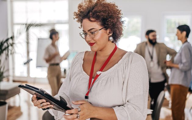A woman in glasses wearing a red lanyard looks down at a tablet in an office, with colleagues standing in the blurred background.
