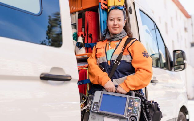 A paramedic in an orange and blue uniform stands by an open ambulance, holding a portable defibrillator while adhering to workforce regulations.