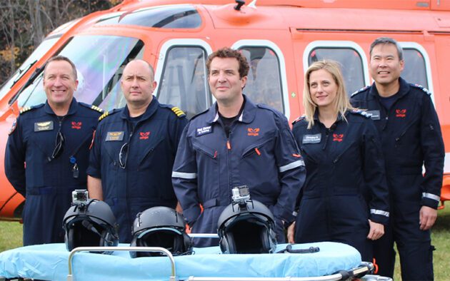 Five Ornge air ambulance crew members in flight suits standing in front of an orange helicopter, with three helmets on a medical stretcher in the foreground.