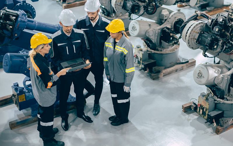 Four factory workers in safety gear stand together in an industrial setting, looking at a laptop computer.