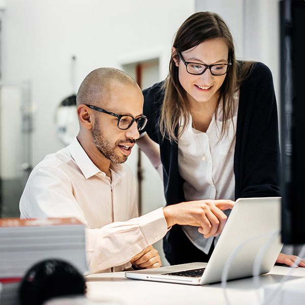 A man and a woman wearing glasses are looking at a laptop together in an office setting. The man is seated and pointing at the screen.