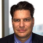 A man with short dark hair wearing a suit and blue shirt sits in front of a window with a blurred background.