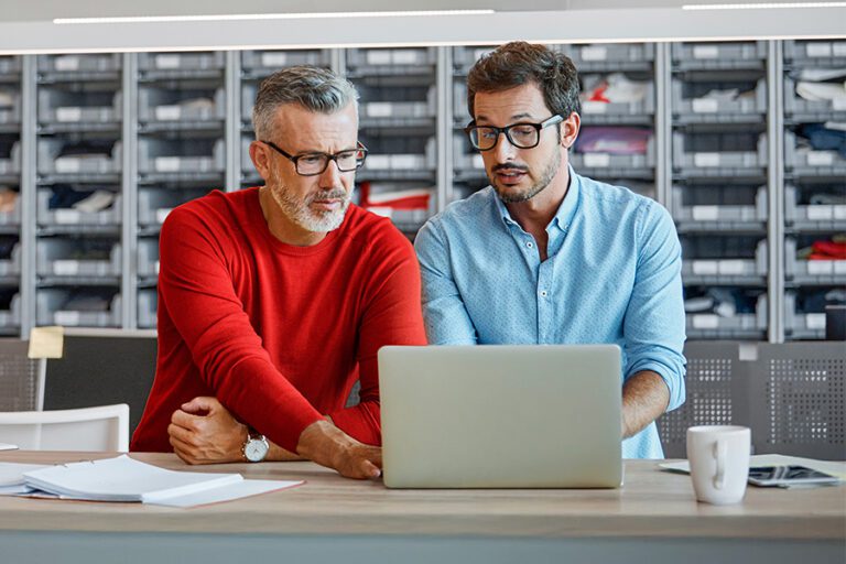 Two men wearing glasses look at a laptop screen together in an office setting, with shelves full of organized boxes in the background.